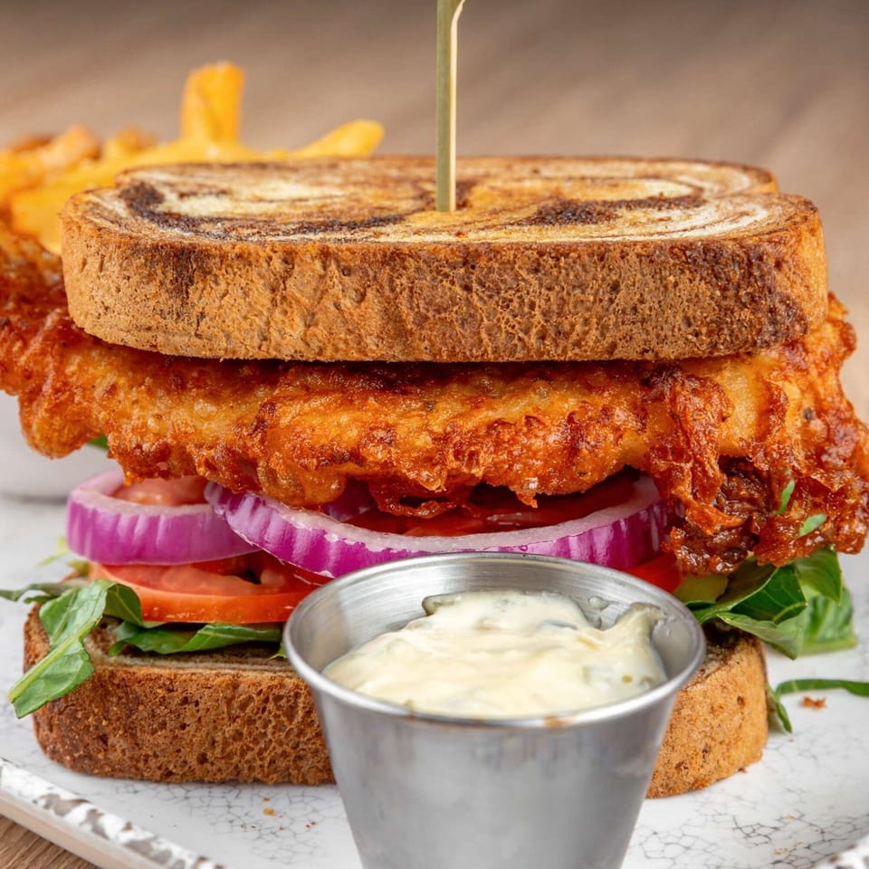 A fried fish sandwich on marbled rye with lettuce, tomato, red onion, and a side of tartar sauce.